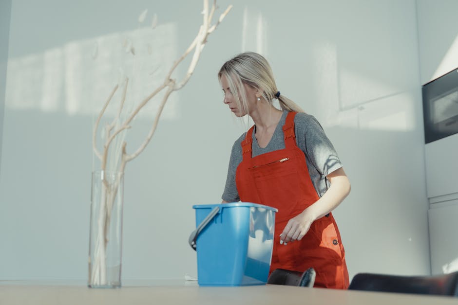 A female cleaning professional from Kingston upon Thames Cleaner stands indoors holding a spray bottle of cleaning solution in one hand and a duster in the other. She is dressed in a black cleaning un