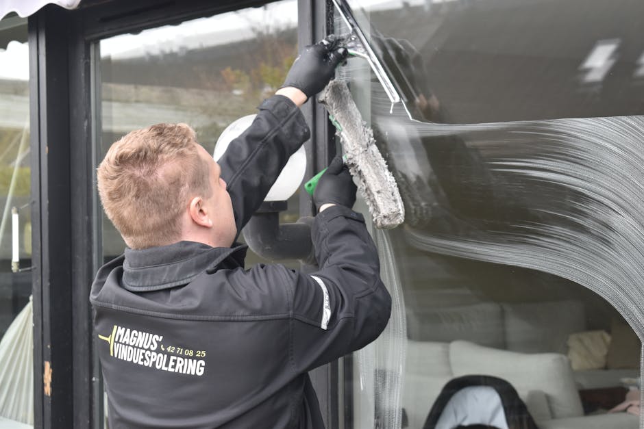 A professional cleaner wearing black gloves and a black jacket with the logo 'Magnus VINDUESPOLERING' on the back is using a long-handled squeegee to clean a large glass window on a commercial bus or 