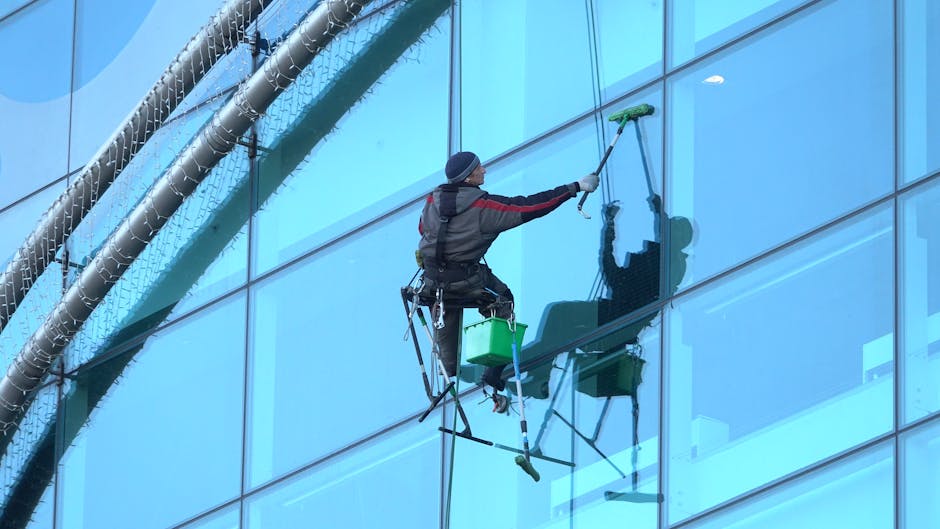 A professional cleaner wearing black gloves and a black jacket with the logo 'Magnus VINDUESPOLERING' on the back is using a long-handled squeegee to clean a large glass window on a commercial bus or 
