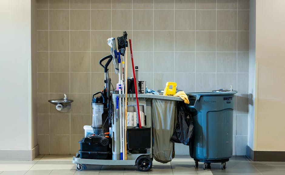 A cleaning trolley positioned against a beige tiled wall in a well-lit room, containing various cleaning tools such as mops, spray bottles, cloths, and a vacuum cleaner. To the left, there is a small