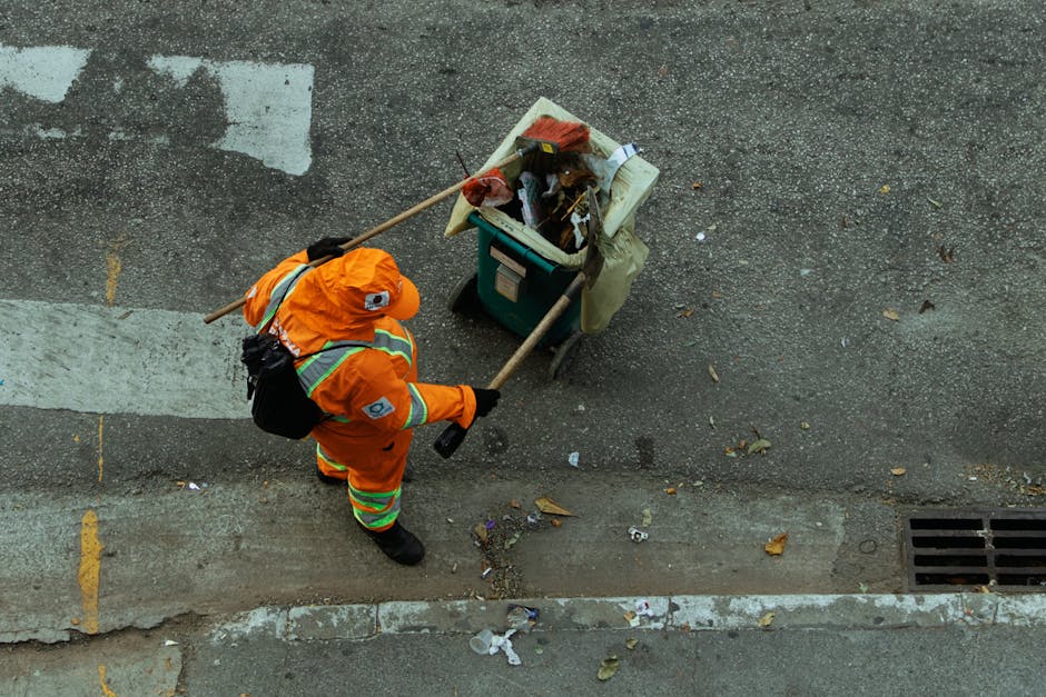 A street cleaner dressed in bright orange high-visibility workwear, including a jacket, trousers, and a cap, is seen from above sweeping a paved sidewalk with a long-handled broom. The cleaner is wear