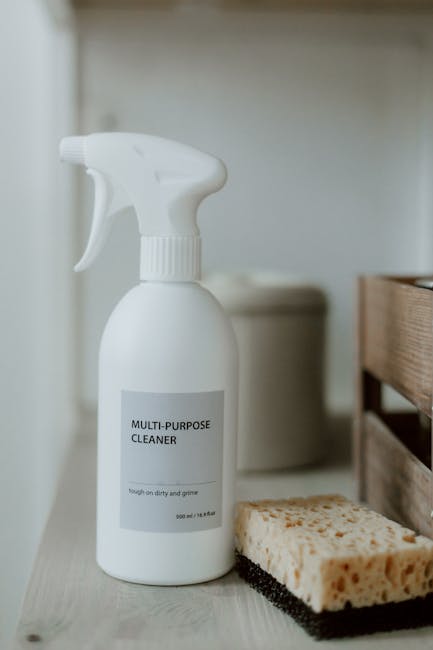 A white plastic spray bottle labeled 'Multi-Purpose Cleaner' is placed on a light-colored kitchen or bathroom countertop alongside a scrubbing sponge with a black abrasive side and a beige porous side, indicating surface cleaning products used for domestic cleaning tasks. In the background, there is a cream-colored container, possibly a waste bin or storage unit, and wooden cabinetry, all set in a well-lit space with neutral tones. The scene reflects a tidy environment prepared for routine or deep cleaning, consistent with the cleaning services offered by Kingston upon Thames Cleaner, as highlighted in the page titled 'KT1 domestic cleaning tips for Kingston Bridge homes'.