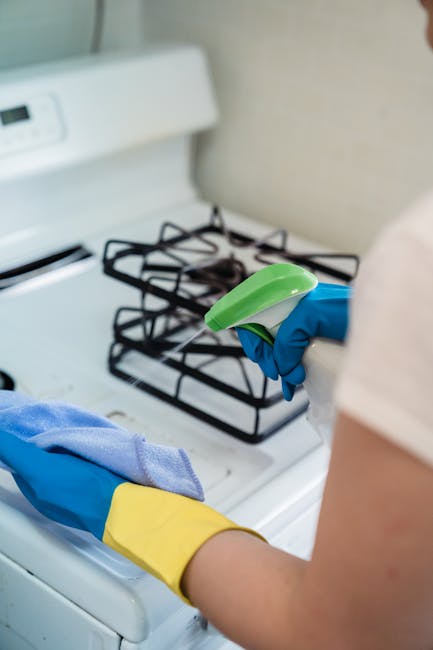A person wearing yellow and blue rubber cleaning gloves is seen holding a spray bottle and a blue cloth while cleaning the surface of a white gas stove in a kitchen. The stove features black metal grates and ceramic burners, with a bright, well-lit environment emphasizing cleanliness and maintenance. The background shows a plain beige wall, and the image highlights surface cleaning and sanitisation processes. This cleaning activity is part of general domestic cleaning services provided by Kingston upon Thames Cleaner, focusing on kitchen hygiene and deep cleaning for residential spaces.