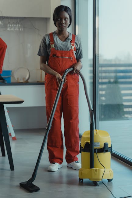 A person wearing white protective coveralls, orange gloves, and white sneakers is operating a yellow and black vacuum cleaner in a modern living room. The room features large floor-to-ceiling glass wi